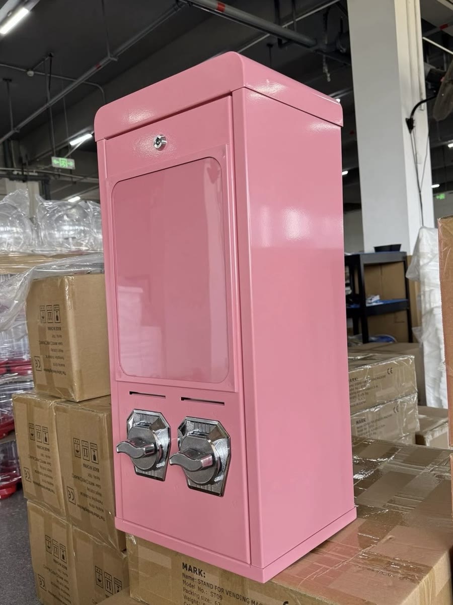 Pinker Postkartenautomat V2 mit zwei Drehknöpfen auf gestapelten Kartons in der Produktionshalle