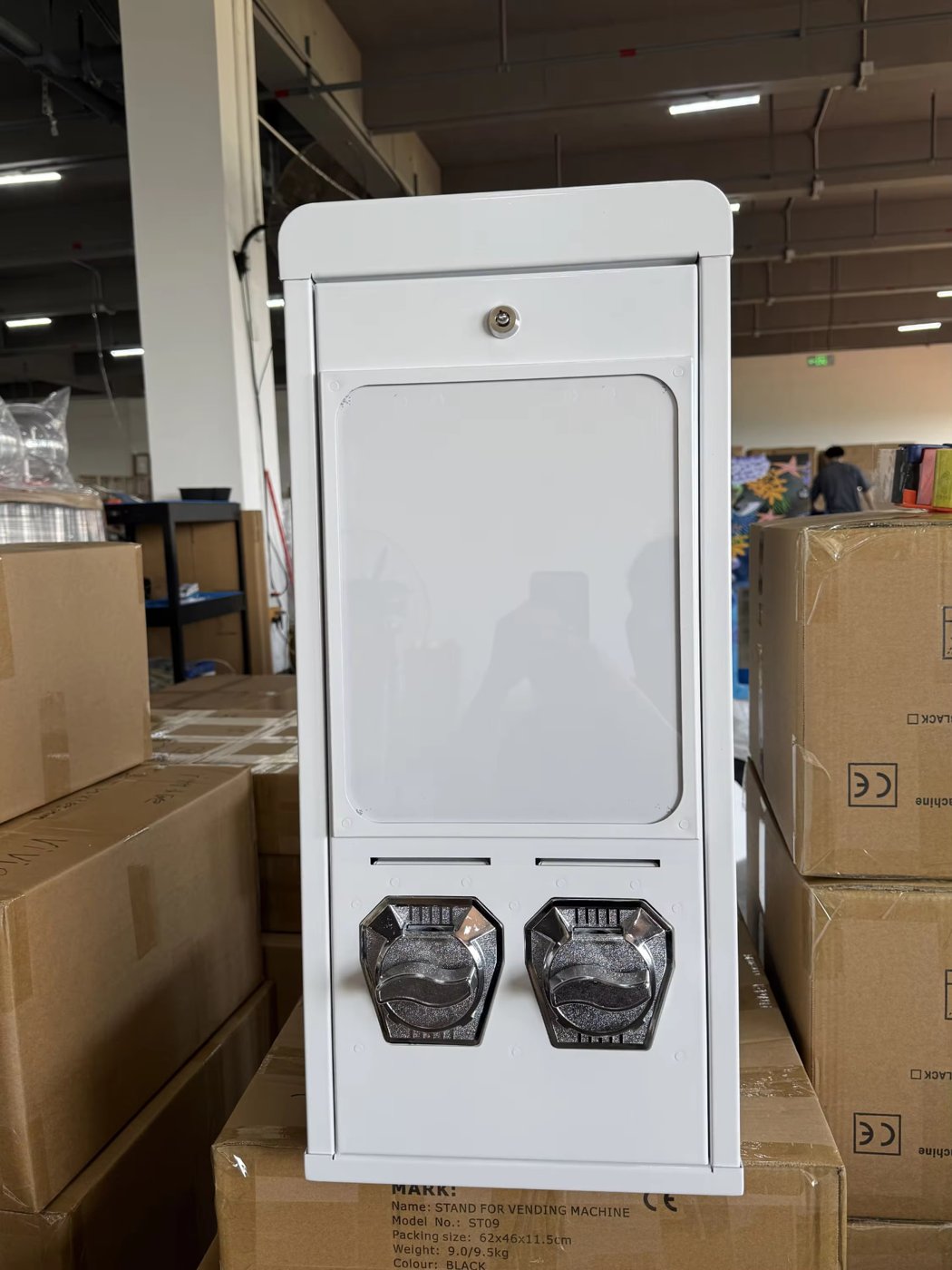 Weißer Postkartenautomat V2 mit zwei Drehknöpfen zwischen Kartons in der Produktion
